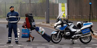 Ein Polizist mit Motorrad sperrt eine Straße ab, dahinter eine Frau mit viel Gepäck