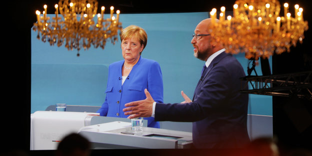 Auf einer Leinwand sind Merkel und Schulz zu sehen, vor ihnen hängen Kronleuchter