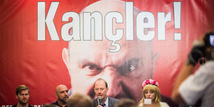 Auf einem roten Plakat steht „Kancler!“, davor sitzen Menschen