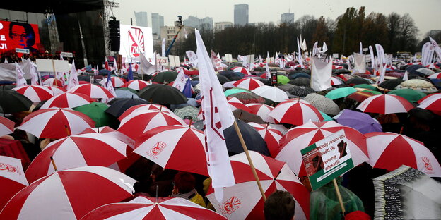 Menschen mit rot-weißen Regenschirmen stehen auf einem Platz.
