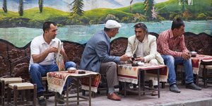 Vier türkische Männer sitzen auf einer Bank vor einer Panorama-Tapete, einer blättert in einer Zeitung, zwei unterhalten sich