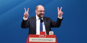 Martin Schulz vor blauem Hintergrund, hinter einem Rednerpult. Er hält die Hände hoch