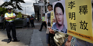 Ldeute halten auf dem Bürgersteig ein Transparent mit dem Kopf von Lee Ming-che, Daneben ein Polizist