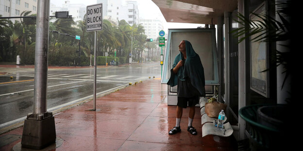 Ein Mann an einer überdachten Bushaltesgtelle, um ihn herum tobt der Hurrican Irma durch die Straßen