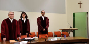 Drei Menschen in Robe stehen hinter einem Tisch in einem Gerichtsaal - oben rechts hängt ein Kreuz an der Wand