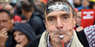 Ein Mann mit Trillerpfeife im Mund und einem "Merkel muss weg"-Aufkleber auf der Stirn, dahinter mehr Menschen