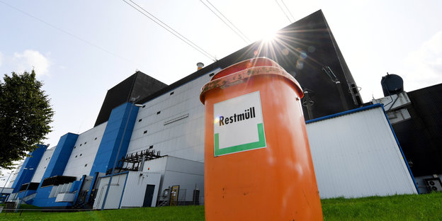Vor einem Atomkraftwerk steht eine orangefarbene Mülltonne, auf der „Restmüll“ steht