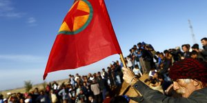 Ein Mann hält eine Fahne der YPG in die Höhe, hinter ihm eine Menschenmenge