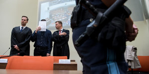 Ein Mann hält ein Papier vor sein Gesicht, neben ihm stehen zwei Männer, ein davon in einer Robe. Im Vordergrund steht ein Justizbeamter