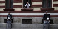Ein Mann guckt aus dem Fenster. Auf dem Bürgersteig stehen zwei Polizisten an einer Hauswand