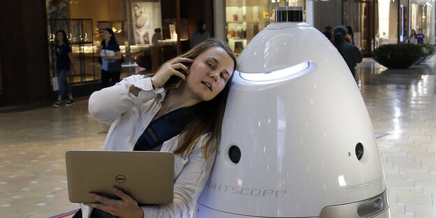 Eine Frau mit einem Laptop in der Hand lehnt sich an einen Roboter Eine Frau mit einem Laptop in der Hand lehnt sich an einen Roboter