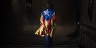 Eine Frau geht eine U-Bahn-Treppe hinunter, es ist sehr dunkel, aber das Geld, Rot und Blau der katalonischen Flagge, die die Frau um den Rücken trägt, ist hell.