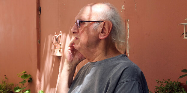 Ein alter Mann mit Halbglatze von der Seite steht vor einem orange-braunem Haus