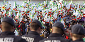 Im Vordergrund sind vier Polizisten von hinten zu sehen, im Hintergrund eine Menschenmenge, die helle Fahnen schwingt