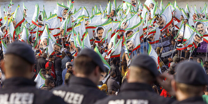 Im Vordergrund sind vier Polizisten von hinten zu sehen, im Hintergrund eine Menschenmenge, die helle Fahnen schwingt