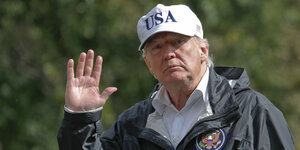 Donald Trump, winkend, mit einer Mütze, auf der „USA“ steht