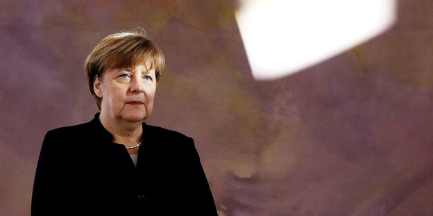 Angela Merkel steht, mit gefalteten Händen und im schwarzen Jackett, vor einem taupe-farbenen Kunstwerk