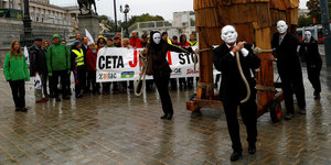 Protest gegen Ceta, Menschen mit Bannern und Menschen mit Masken, die ein Holzpferd ziehen