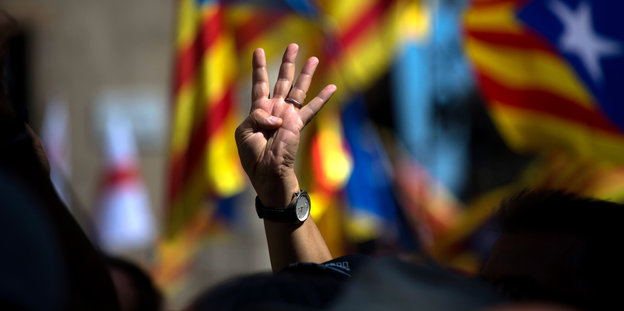 Ein Mensch hält vier Finger in die Höhe. Dahinter sieht man Estelada-Flaggen, offizielle Flaggen der autonomen spanischen Region Katalonien