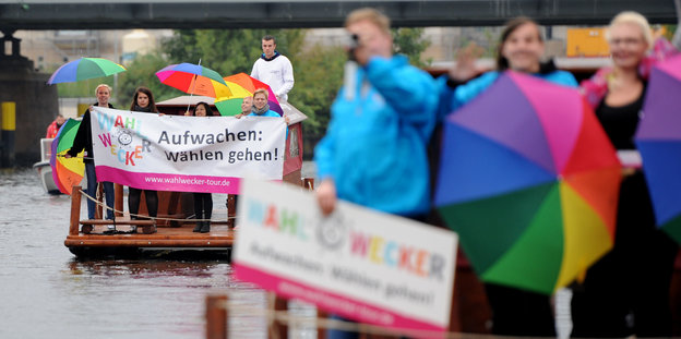 Menschen auf Flößen in einem Fluß, sie tragen bunte Regenschirme und ein Transparent mit der Aufschrift "Aufwachen: Wählen gehen"