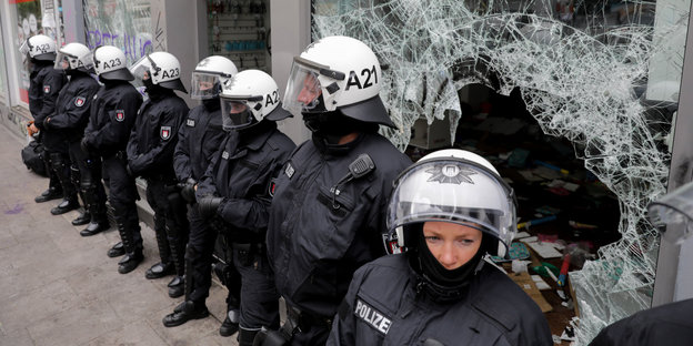 Polizisten in Schutzkleidung und mit Helmen stehen vor einer zerstörten Glasscheibe in einer Reihe nebeneinander
