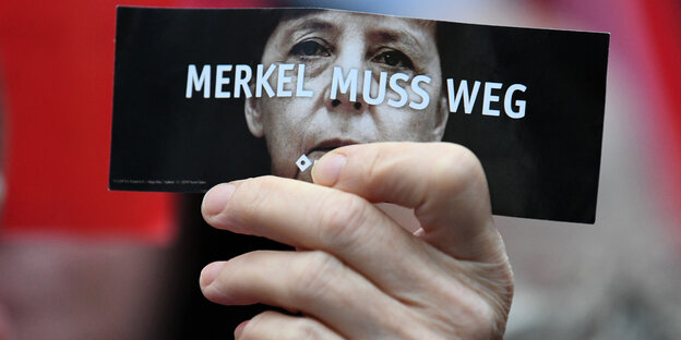 Eine Hand hält einen Flyer mit dem Gesicht von Angela Merkel und dem Spruch "Merkel muss weg"