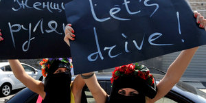 Zwei Femen-Aktivistinnen mit nackten Oberkörpern und vermummten Gesichtern halten Schilder hoch, auf denen "Let's drive" steht
