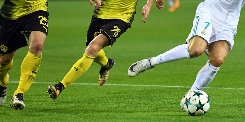 Blick auf die Beine bei einem Fußballspiel. Drei Personen versuchen den Ball zu kriegen