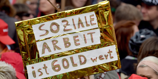 Auf einer Demonstration wird ein goldenes Schild mit der Aufschrift "Sozial Arbeit ist Gold wert" hochgehalten.