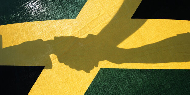 Die Schatten von drei Händen, die einander halten. Davor eine Jamaika-Flagge.