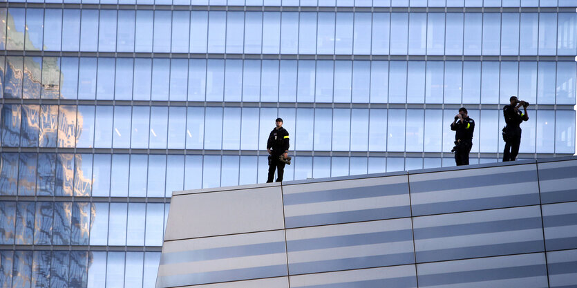 Drei Polizeibeamte stehen auf einem spiegelnden Haus, im Hintergrund ein höheres, ebenfalls spiegelndes Haus