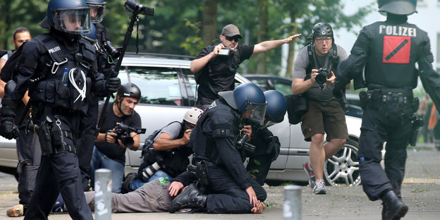 Polizisten drücken einen Mann auf den Boden, ein Mann im Hintergrund hält einen Fotoapparat