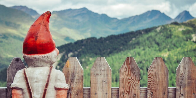 Rückenansicht eines Gartenzwergs, der an einem Zaun steht und Berge betrachtet