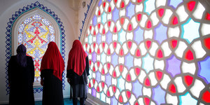 Drei Frauen stehen, dem Betrachter abgewandt vor einem leuchtenden, rot und türkis gefärbten Glasmosaik, zwei von ihnen tragen rote Kopftücher