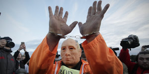 Ein Mann mit einer Putin-Maske hebt beide Hände die mit Handschellen verbunden sind