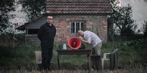 Zwei Männer stehen in einem Garten und lauschen an einem Trichter