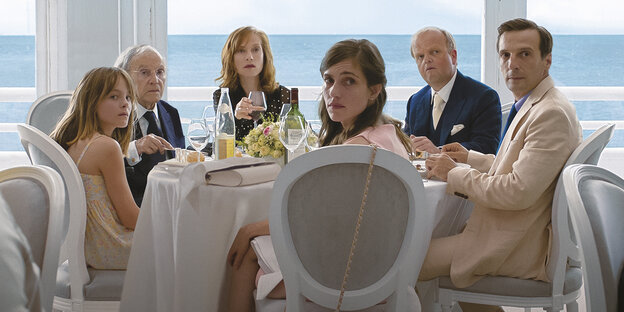 An einem Tisch sitzen 6 Menschen, unterschiedlichen Alters, vornehm gekleidet, vor einer Meereskulisse