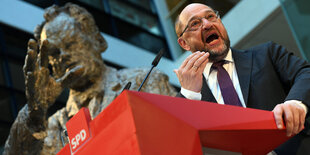 Ein Mann steht auf einem Podium und gestikuliert. Hinter ihm ist eine Statue zu sehen