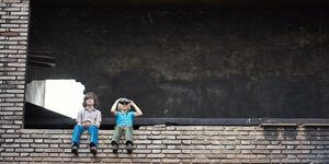 Zwei Kinder sitzen nebeneinander auf der Kante einer Mauer, der Kleinere guckt durch ein Fernglas