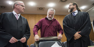 Drei Männer im Gerichtsaal, zwei Anwälte und in ihrer Mitte der Angeklagte
