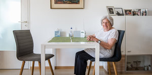 Eine ältere Dame sitzt an einem Tisch in ihrer Wohnung und blickt in Richtung Kamera