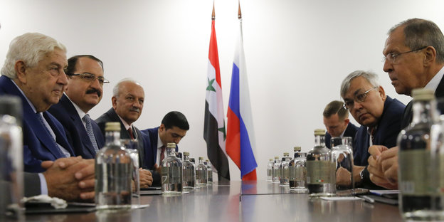 Viele Männer sitzen an einem langen Tisch. Im Hintergrund eine russische und eine syrische Flagge