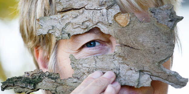 Eine Frau hält sich ein Stück Baumrinde vors Gesicht, ein Auge ist frei