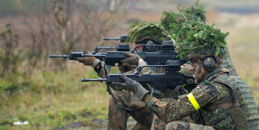 Mehrere Soldaten knien in einer Reihe mit angelegtem Gewehr und Tarnhüten.