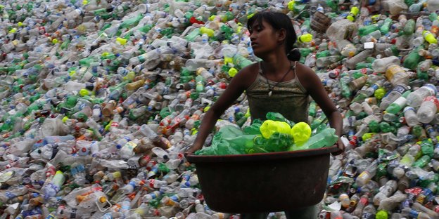 Mädchen trägt Korb voller Plastikflaschen, dahinter ein riesiger Haufen Flaschen
