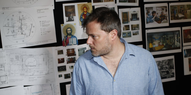 Ein Mann in blauem Hemd vor einer Wand, an der viele kleine Bilder aufgehängt sind