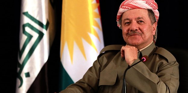 Ein Mann sitzt vor zwei Flaggen und lächelt