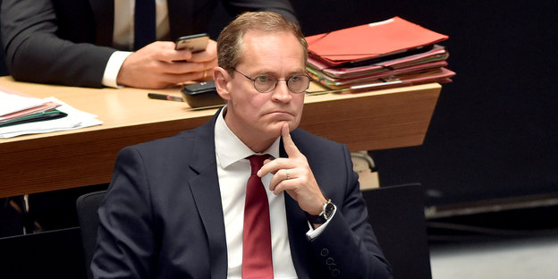 Mann mit Brille sitzt im Anzug im Parlament, kneift das Gesicht zusammen