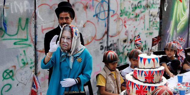 Eine Frau als Queen Elizabeth II verkleidet, geht winkend an einer Mauer vorbei