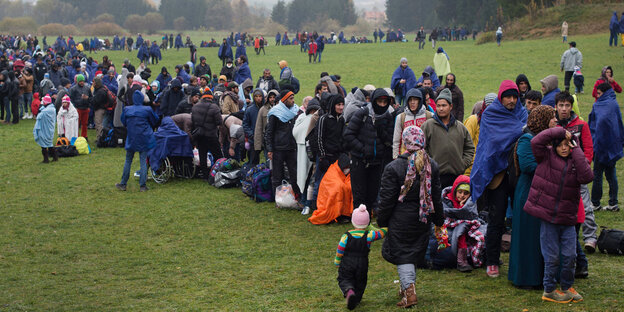 Flüchtlinge in mehr oder weniger warmer Kleidung stehen auf einer kalt und verregnet aussehenden Wiese